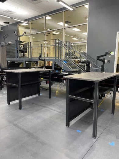 Steel-frame work tables with woodgrain tops and monitor arms beside a platform with cable railing