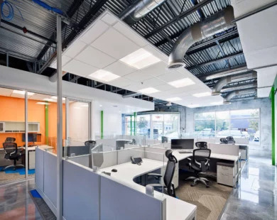 Corner workstations with glass screens and black ergonomic chairs near an orange-walled glass room