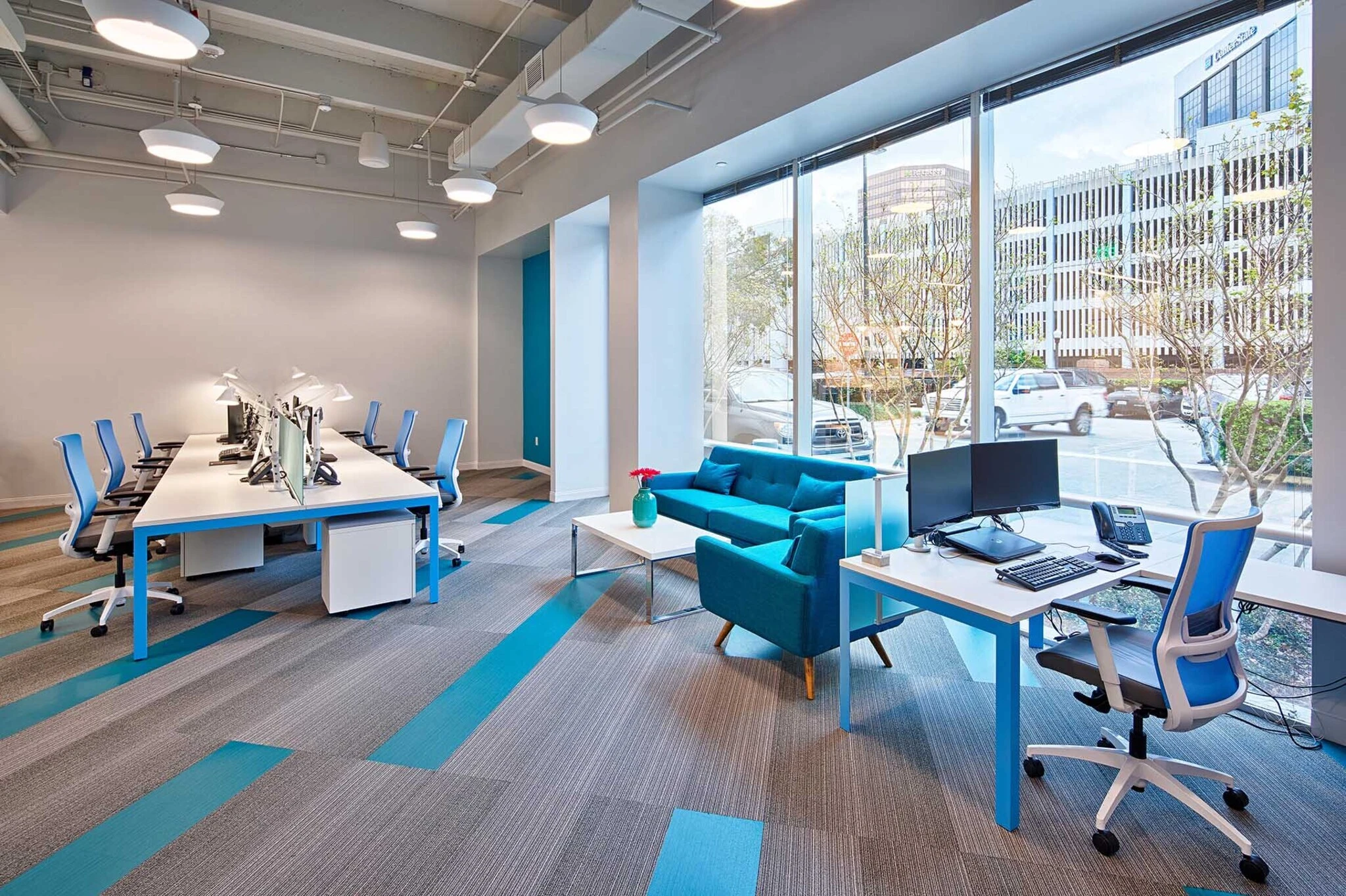 Open office benching workstations with blue ergonomic task chairs and a blue lounge seating area beside floor-to-ceiling windows