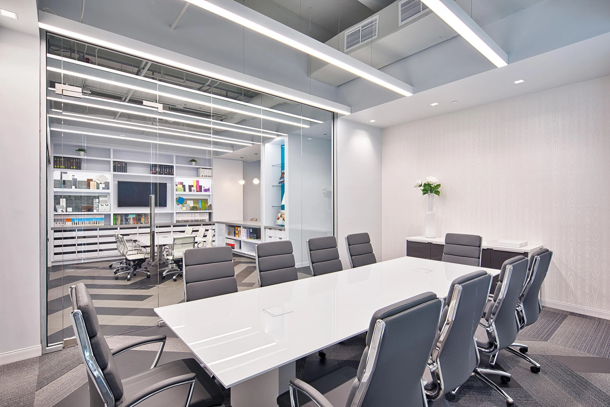 Ten-seat boardroom with white conference table and gray executive chairs, with the material library visible through glass