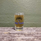 A clear Uranus Fudge Factory rocket logo shot glass filled with a bubbly drink against a textured green background