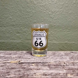 A collectible Uranus US 66 highway route shield shot glass filled with cider on a rustic wood plank background
