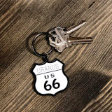 A black-and-white metal keychain shaped like a classic highway shield reading "URANUS US 66", attached to a keyring with two silver house keys and resting on a textured dark wood background.