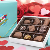 Blue gift box with nine assorted Uranus chocolates displayed on a table with hearts and roses in front of a heart background for Valentine's Day.