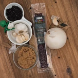 A "Jerk Uranus" Garlic & Black Pepper Beef Strip is centered on a rustic wood surface, surrounded by its key ingredients: a bowl of black peppercorns, a white bowl of garlic cloves, a whole white onion, a dish of brown sugar, and a small green alien figurine peeking out from the side.