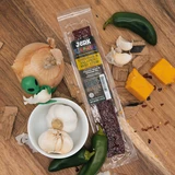 A "Jerk Uranus" Cheddar & Jalapeño Beef Strip is displayed on a rustic wood surface, surrounded by fresh ingredients: whole garlic bulbs in a white bowl, a yellow onion, fresh green jalapeños, blocks of sharp cheddar cheese, and a tiny green alien figurine peeking out from behind the onion.