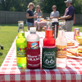 Outdoor picnic scene with Uranus sodas in red and green koozies on a red-and-white checkered tablecloth, featuring “Show Me Uranus” and “Uranus US 66” designs with people socializing in the background.