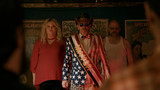 Three characters stand under dim carnival lights — a man in a red, white, and blue “Mayor” suit with sunglasses, flanked by two serious companions — in a tense scene from “Uranus Attacks.”
