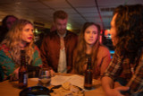 A group of friends sit around a bar table with drinks, debating a plan in a small-town dive bar during a scene from “Uranus Attacks.”