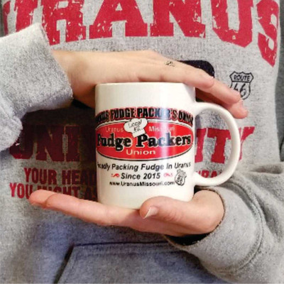A pair of hands holds a white ceramic coffee mug in front of a person wearing a heather grey "Uranus" sweatshirt. The mug features a red oval logo for the "Uranus Fudge Packers Union" complete with a chef's hat reading "Local #2", followed by black text below that boasts: "Proudly Packing Fudge In Uranus Since 2015".