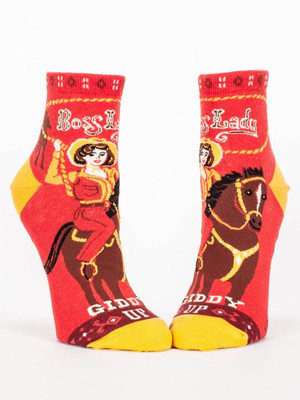 A pair of bright red ankle socks displayed against a plain white background. The socks feature mustard yellow accents on the toe and heel, along with dark burgundy western-patterned bands on the top cuff and near the toe. The main graphic is a vintage illustration of a cowgirl wearing a yellow hat and shirt, swinging a yellow lasso while riding a brown horse. Framed by a yellow rope design at the top, decorative white text reads: "Boss Lady". Down near the toe, white western-style text finishes with: "GIDDY UP".