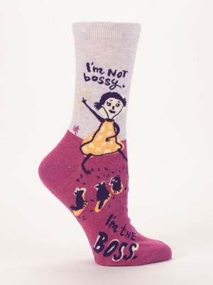 A single crew sock displayed against a plain white background. The upper half of the sock is light heather grey, while the lower foot section, including the heel and toe, is a heathered magenta pink. Knit into the side is an illustration of a confident girl in a yellow dress pointing her arm outward, with three small birds following at her feet. Above the girl on the grey background, dark text reads, "I'm Not bossy." Down by the toe on the pink background, the text finishes with: "I'm the BOSS."