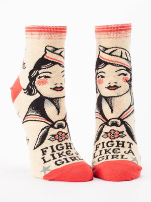 A pair of light cream ankle socks displayed against a plain white background. The socks feature coral-red accents on the toe, heel, and striped top cuff. The main design is a vintage, traditional tattoo-style illustration of a smiling sailor girl with black hair, a white sailor hat, and rosy coral cheeks. Below her sailor collar is a small traditional tattoo-style flower. Near the toe, bold black retro text reads: "FIGHT LIKE A GIRL", accented by a few scattered grey nautical stars.