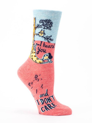 A single crew sock displayed against a plain white background. The top third of the sock is light blue, while the bottom two-thirds, including the heel and toe, are a heathered coral pink. Knit into the side is an illustration of a tree with a small owl perched on a branch. Below the owl, a person is relaxing in a yellow hammock tied to the tree trunk. Dark cursive text next to the hammock reads, "I heard you". Further down the foot, bold dark text finishes the thought: "and I DON'T CARE".