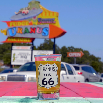 A Uranus US 66 shield shot glass filled with amber liquid, with the Uranus Fudge Factory sign background