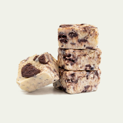 A stack of three square pieces of cookies and cream fudge embedded with chunks of chocolate sandwich cookies, with a fourth piece leaning against the base, set against a solid off-white background.