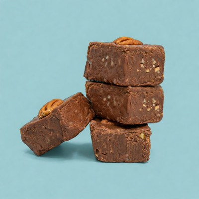A stack of three square pieces of chocolate pecan fudge studded with chopped nuts and topped with whole pecans, with a fourth piece leaning against the base, set against a solid light blue background.