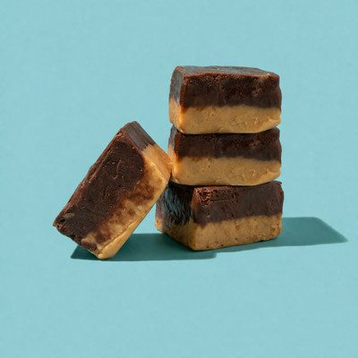 A stack of three square pieces of two-layered chocolate and peanut butter fudge, with a fourth piece leaning against the base, set against a solid light blue background.