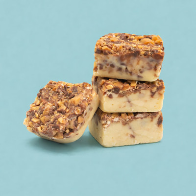 A stack of three square pieces of toffee fudge topped with crushed toffee and chocolate bits, with a fourth piece leaning against the base, set against a solid light blue background.