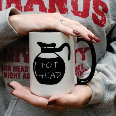 A pair of hands with dark red nail polish holds a white ceramic coffee mug with a black handle in front of a person's chest. Printed on the front of the mug is a black silhouette of a classic diner-style coffee pot. Inside the black coffee pot graphic, stacked white handwritten-style text reads "POT HEAD". The person in the background is wearing a heather grey sweatshirt with distressed red text that partially reads "URANUS".