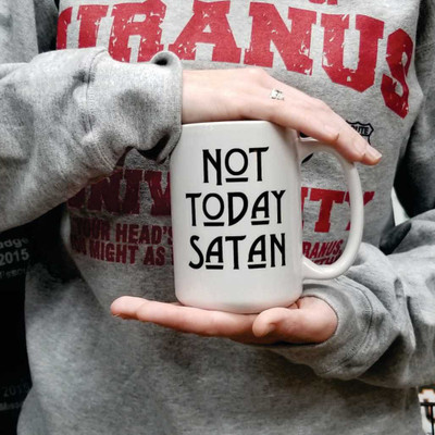 A pair of hands holds a white ceramic coffee mug in front of a person's chest. Printed on the front of the mug is the stacked, black text "NOT TODAY SATAN" in a stylized, blocky font with horizontal lines underlining the O and the As. The person in the background is wearing a heather grey sweatshirt with distressed red text that partially reads "URANUS UNIVERSITY".