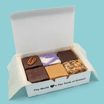 An open white box lined with parchment paper displays a six-piece assortment of diverse fudge squares—including chocolate pecan, purple marbled, plain chocolate, peanut butter, and a loaded nut-and-chip variety—against a light blue background, with the front edge reading "The World loves The Taste of Uranus!".