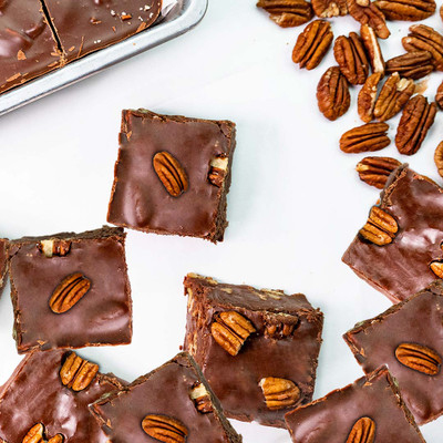 A top-down view of several thick, square pieces of chocolate fudge scattered across a clean white surface. Each piece of fudge has a whole pecan half pressed into the top. A pile of loose pecan halves rests in the upper right corner.