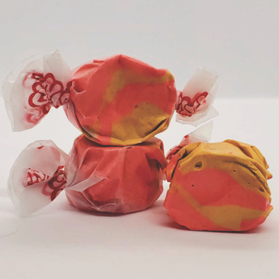 Three pieces of pink and orange swirled saltwater taffy are displayed against a white background, with two pieces stacked and one sitting to the side, all partially wrapped in wax paper with red twisted ends.