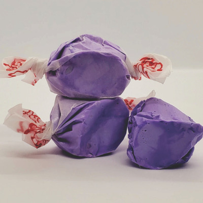 Three pieces of solid purple taffy, wrapped in clear paper with red logos on the twisted ends, are arranged on a plain white surface. Two pieces are stacked on top of each other, while the third piece rests against them on the right.