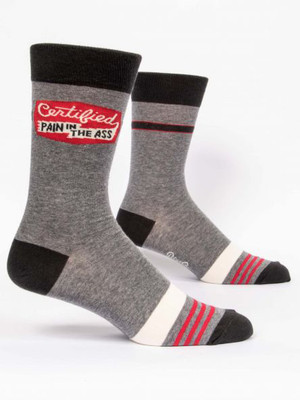 A pair of heather grey crew socks displayed against a plain white background. The socks feature black accents on the toe, heel, and top cuff. Across the mid-foot is a wide cream-colored stripe followed by a section with three thin red stripes. Knit into the upper ankle of the front sock is a distressed red graphic resembling a stamped badge. Inside the badge, white cursive text reads "Certified" above a white banner containing the black block text: "PAIN IN THE ASS".