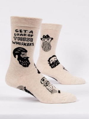 A pair of light heather tan crew socks displayed against a plain white background. The socks are decorated with a scattered pattern of various cartoon men's faces featuring large, bushy beards and mustaches drawn in black, grey, and white. Knit into the upper ankle of the front sock is the bold, stacked black text: "GET A LOAD OF THESE WHISKERS".