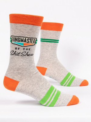 A pair of light heather grey crew socks displayed against a plain white background. The socks feature bright orange accents on the toe, heel, and top cuff, along with two thin green stripes on the upper calf and three thicker lime green stripes across the foot. Knit into the ankle area of the front sock is a retro-style graphic with the word "RINGMASTER" in tall black letters inside an outlined box, underlined by geometric teal and orange shapes. Below the graphic, black text reads "OF THE" in small caps, followed by "Shit Show" in a bold cursive script.