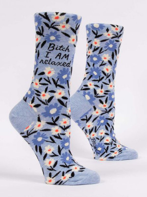 A pair of light blue crew socks displayed against a plain white background. The socks feature an all-over floral pattern with medium blue and white flowers, orange centers, and black stems and leaves. Woven into the upper ankle of the front sock is the black script text: "Bitch I AM relaxed".