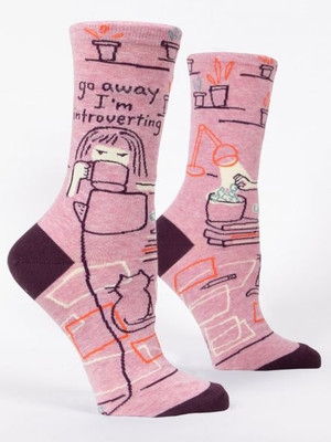 A pair of heathered pink crew socks displayed against a plain white background. The socks feature dark plum accents on the toe and heel. They are decorated with sketchy line-art illustrations of a cozy room featuring shelves of potted plants, a desk lamp shining on a bowl of snacks, stacked books, and a cat sitting on a geometric rug. On the ankle of the front sock, a girl wrapped in a blanket glares over a mug she is drinking from. Above her head, dark text reads: "go away I'm introverting".