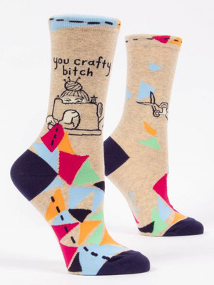 A pair of light tan crew socks displayed against a plain white background. The socks feature dark navy blue accents on the toe and heel, and a light blue top cuff detailed with black dashed lines resembling stitches. The foot of the sock is covered in a vibrant geometric patchwork pattern of pink, orange, light blue, mint green, and navy triangles connected by dashed stitch lines. On the ankle of the front sock is a line-art illustration of a woman with knitting needles in her hair bun, peering over a sewing machine as she stitches the patchwork together. Above her head, dark handwritten-style text reads: "you crafty bitch".