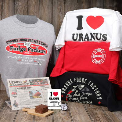 A sprawling Uranus souvenir bundle displayed on a rustic wooden background, featuring a folded stack of four cheeky graphic tees—including "I Heart Uranus" and "Fudge Packer's Union"—alongside a local newspaper, a matching sticker, a Route 66 keychain, and a thick square of fudge resting on a mini cutting board.