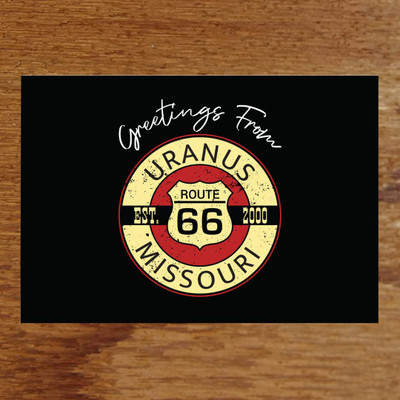 A horizontal postcard featuring a solid black background with white cursive text reading "Greetings From" centered at the top. Below is a distressed, circular vintage-style emblem with a Route 66 shield in the red center, framed by a yellow ring reading "URANUS MISSOURI" and crossed by a black banner reading "EST. 2000".