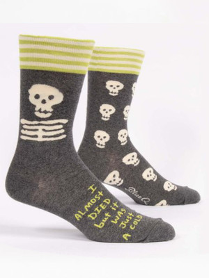 A pair of dark heather grey crew socks displayed against a plain white background. The socks feature a top cuff striped in lime green and cream. The front sock displays a large cream-colored cartoon skull and ribcage on the ankle. Knit into the side of the foot below it is bright lime green text that reads: "I ALMOST DIED but it WAS Just A COLD". The back sock shows that the rest of the design features an all-over pattern of smaller cream skulls.