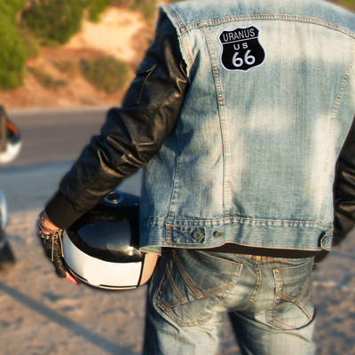 Person wearing a denim and leather motorcycle jacket with a Uranus Route 66 embroidered patch on the back, holding a helmet while standing outdoors near a road.