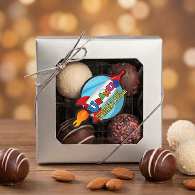 Assorted gourmet chocolate truffles in a 4-piece silver gift box from Uranus Fudge Factory, including a variety of milk, dark, and white chocolate truffles with decorative drizzles and textured coatings, displayed with almonds on a wooden surface.