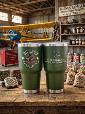 Green Uranus Crop Dusters insulated tumbler with vintage airplane design displayed in rustic hangar setting with yellow crop duster plane and gift shop items.
