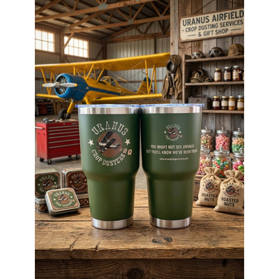 Green Uranus Crop Dusters insulated tumbler with vintage airplane design displayed in rustic hangar setting with yellow crop duster plane and gift shop items.
