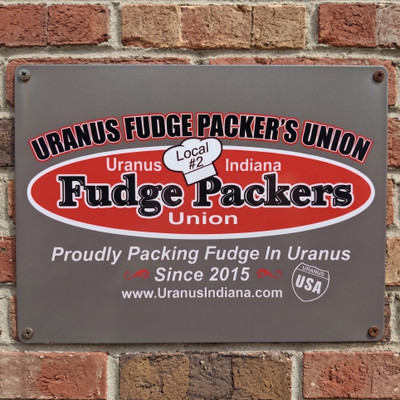 Uranus Fudge Packer's Union 12x16 metal sign mounted on a red brick wall, featuring the red and white Fudge Packers Union oval logo, Local #2 chef's hat, and "Proudly Packing Fudge In Uranus Since 2015" tagline.