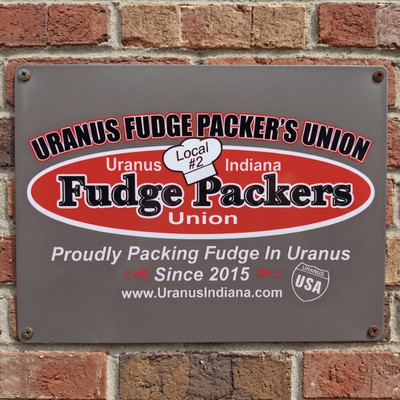 Uranus Fudge Packer's Union 12x16 metal sign mounted on a red brick wall, featuring the red and white Fudge Packers Union oval logo, Local #2 chef's hat, and "Proudly Packing Fudge In Uranus Since 2015" tagline.