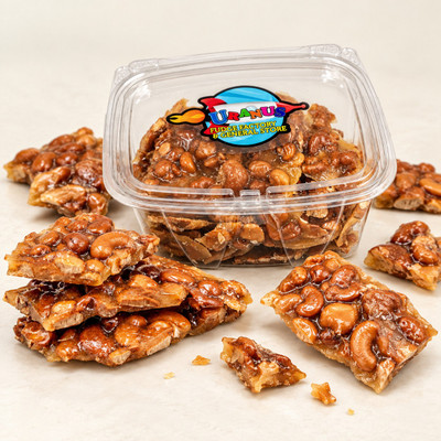 Uranus Fudge Factory cashew brittle in a clear 1 lb. tub with broken pieces of glossy cashew brittle scattered around it