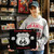 A young woman in a gray hoodie holding up a black Uranus Missouri Route 66 metal souvenir sign inside a shop