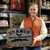 A smiling young woman in an orange quilted vest holding up a novelty Uranus Meat & Sausage Co. metal sign in a souvenir shop