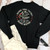 A flat lay of a black crewneck sweatshirt paired with white denim and sunglasses on a white wood background. The sweatshirt features a large circular "Uranus Fudge Factory" logo.