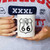 A pair of hands holds a white ceramic coffee mug in front of a person wearing a heather grey graphic shirt. Printed boldly on the center of the mug is a classic, black-and-white Route US 66 highway shield.
