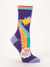 A single dark purple crew sock displayed against a plain white background. The sock features light blue accents on the toe and heel, and a rainbow-striped top cuff. Knit into the side is a small, round yellow cartoon character shooting a colorful rainbow from its backside down into a fluffy white cloud.